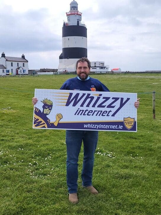 James O'Sullivan, owner of Whizzy Internet, standing in front of Hook Lighthouse in County Wexford with a sign with the Whizzy logo on it as well as Mr. Whizzy to signify that Whizzy Internet broadband is available in this area.